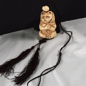 May include: A cream-colored carved netsuke pendant featuring a seated cat with its paws crossed, wearing a hood. The pendant is attached to a dark brown cord with two long, matching tassels hanging from the bottom and a small circular bead at the top.