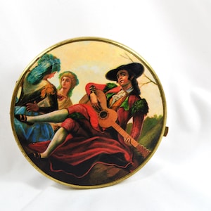 May include: A round, vintage compact with a painted scene of a man playing a guitar, surrounded by two women. The man wears a red coat and black hat. The compact has a gold-colored metal frame.