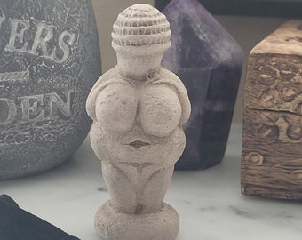 Pode incluir: Uma pequena estatueta de Vénus bege com base arredondada, seios proeminentes e cabeça detalhada. A estátua é exibida com outros itens decorativos, incluindo um vaso de pedra cinza escuro e um cristal roxo.