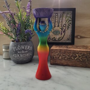 May include: A rainbow colored resin statue of a woman holding a purple bowl above her head. The statue is standing on a white surface.