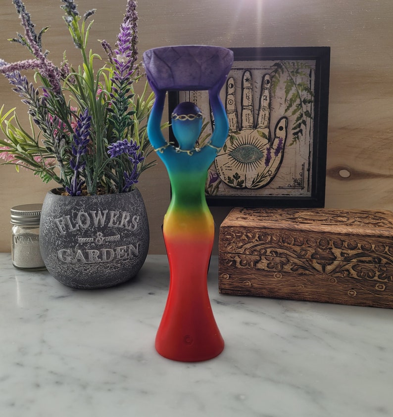 May include: A rainbow colored resin statue of a woman holding a purple bowl above her head. The statue is standing on a white surface.