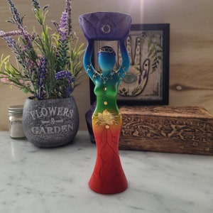 May include: A colorful wooden statue of a woman holding a bowl above her head. The statue is painted in shades of red, orange, yellow, green, blue, and purple. The bowl is purple with a gold circle in the center. The statue is standing on a white surface.