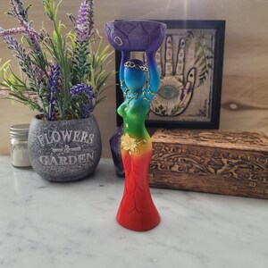 May include: A rainbow colored wooden statue of a woman holding a purple lotus flower above her head. The statue is decorated with gold accents and has a floral design on the skirt.