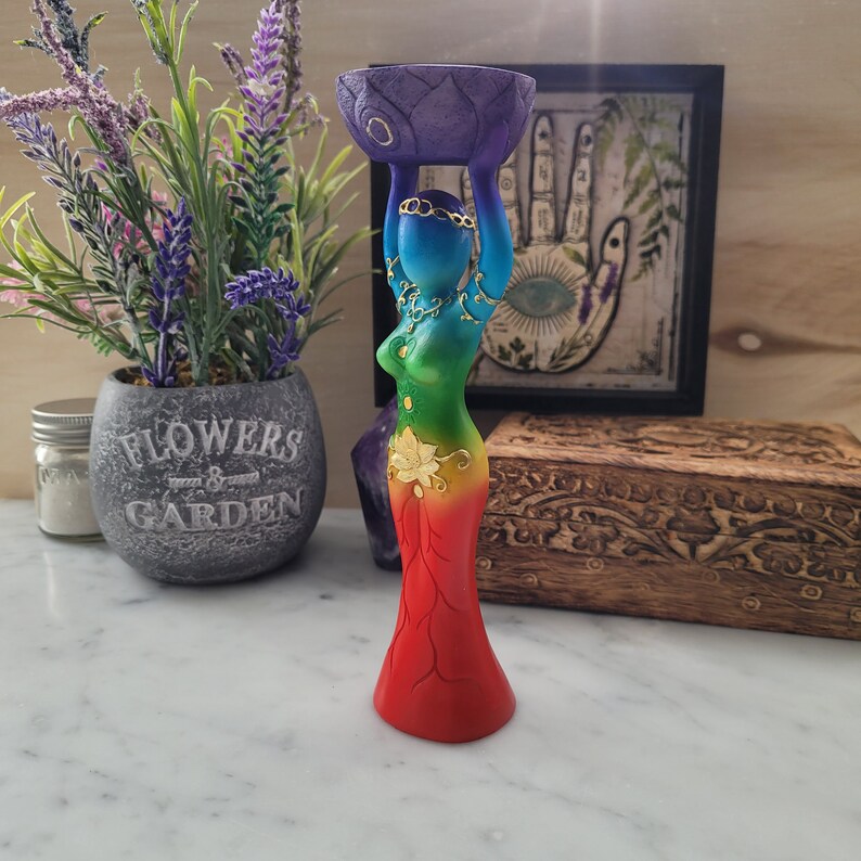 May include: A rainbow colored wooden statue of a woman holding a purple lotus flower above her head. The statue is decorated with gold accents and has a floral design on the skirt.
