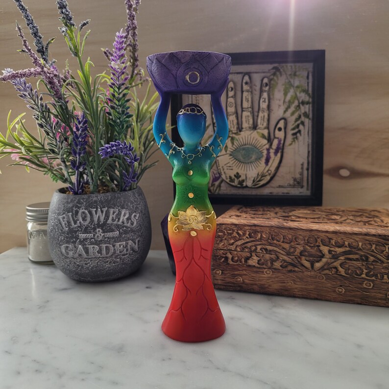 May include: A rainbow colored wooden statue of a woman holding a purple bowl above her head. The statue has gold accents and is standing on a white surface.