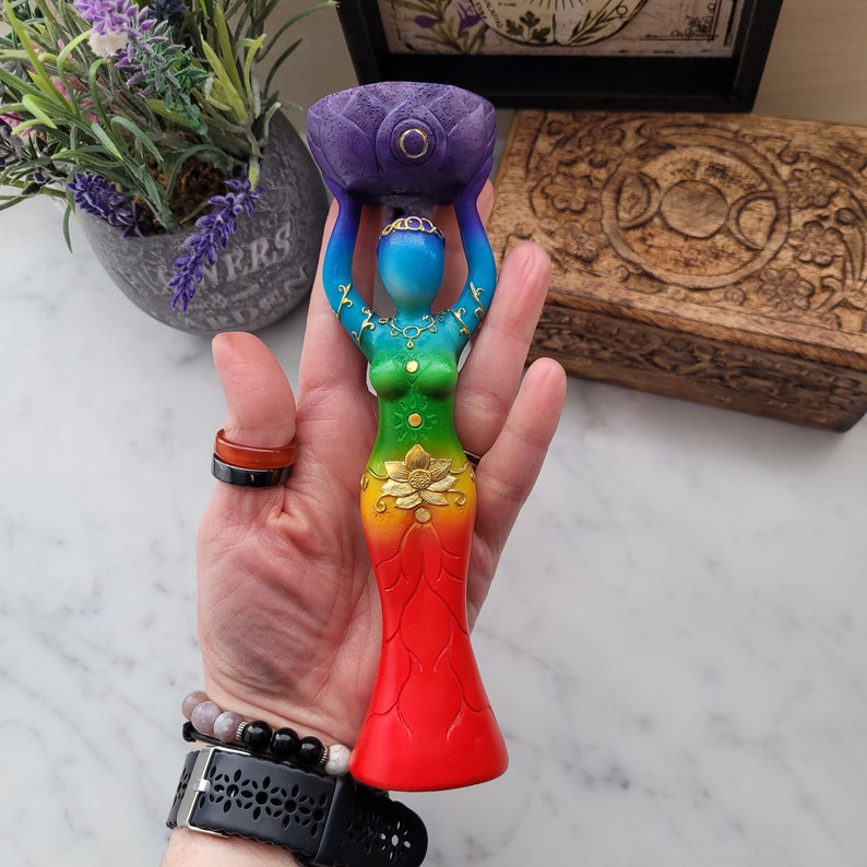 May include: A rainbow colored wooden goddess statue with a lotus flower on her head and a gold lotus flower on her stomach. She is holding a purple lotus flower bowl in her hands.