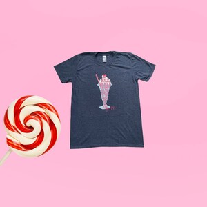 May include: A dark heather grey t-shirt featuring a graphic of a milkshake in a glass with the words "Milk Shake" and "All the things you love" written in pink and white.