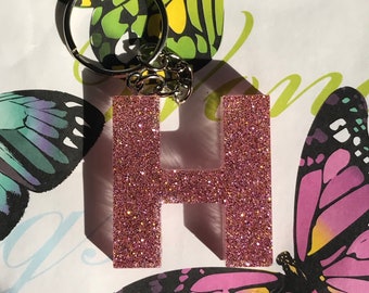 Op de afbeelding: Een roze glitter letter H sleutelhanger met een zilveren sleutelring. De sleutelhanger is op een witte achtergrond met vlinderpatronen.