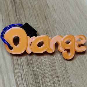 Könnte beinhalten: Orangener Schlüsselanhänger mit einem blauen und roten Band. Das Wort "Orange" ist in orangefarbenem Kunststoff geschrieben.
