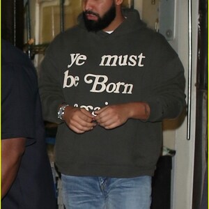 May include: A man wearing a dark green hooded sweatshirt with the text "ye must be Born" in white lettering. He is wearing blue jeans and has a beard.