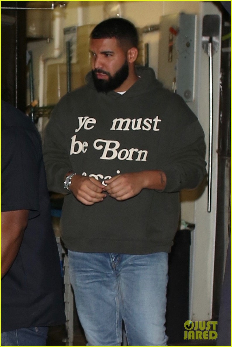 May include: A man wearing a dark green hooded sweatshirt with the text "ye must be Born" in white lettering. He is wearing blue jeans and has a beard.