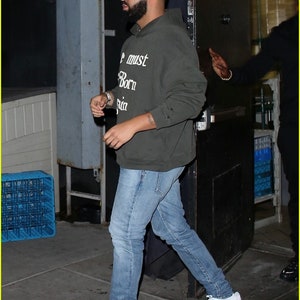 May include: A man wearing a dark green hooded sweatshirt with the text "We must be born again" printed on the front. He is wearing blue jeans and white sneakers.