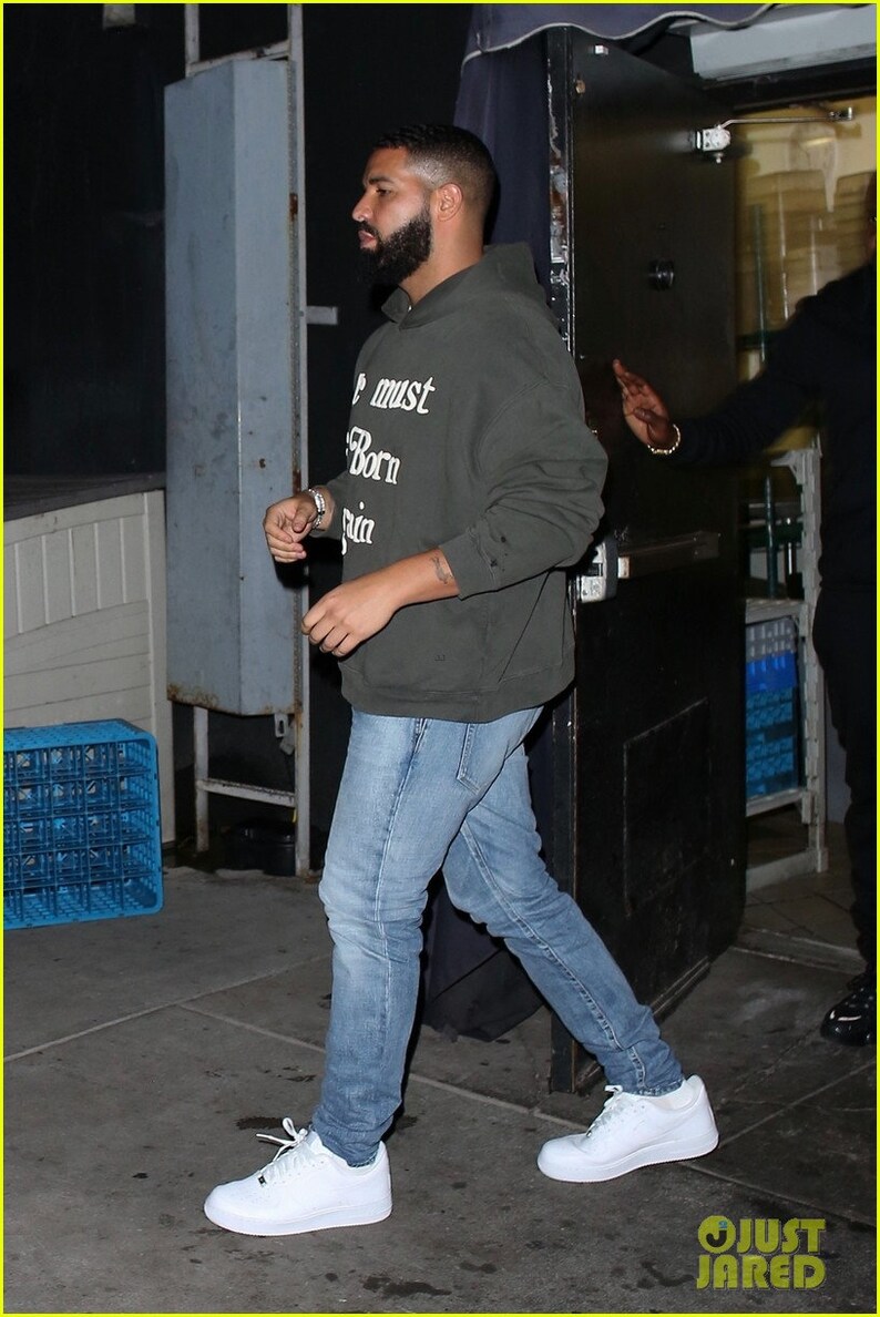 May include: A man wearing a dark green hooded sweatshirt with the text "We must be born again" printed on the front. He is wearing blue jeans and white sneakers.