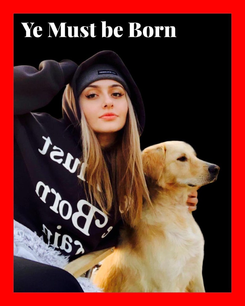May include: A young woman wearing a black hoodie with the text "Ye Must be Born" in white lettering, a black beanie, and a black and white graphic on the front of the hoodie. She is holding a golden retriever puppy in her arms.