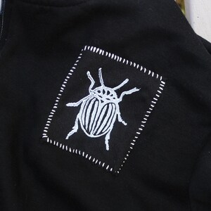 May include: Black zippered hoodie with a white embroidered insect design on the front. The insect is a beetle with a striped body and long legs.