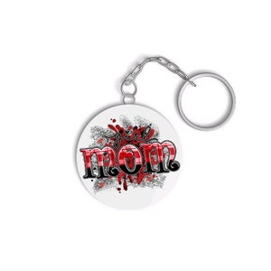 May include: A white round keychain with a red and black plaid design that says "girl mom" in black lettering with a red and black plaid background. The keychain has a silver metal ring.