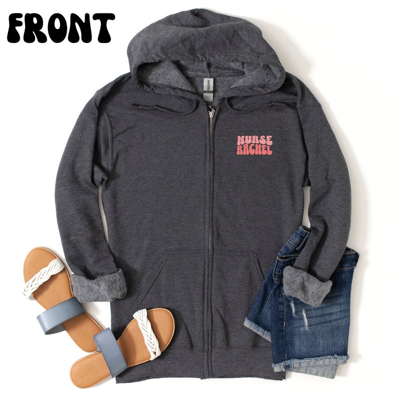 Puede incluir: Una sudadera con capucha gris oscuro con cremallera y el texto "NURSE RACHEL" en rosa. La sudadera con capucha se combina con pantalones cortos de mezclilla y sandalias con correas trenzadas blancas y suelas azules. La palabra "FRONT" est&aacute; en la parte superior de la imagen.