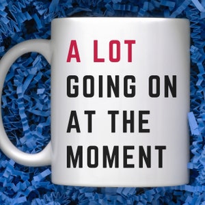 May include: White ceramic coffee mug with black text that reads "A LOT GOING ON AT THE MOMENT". The word "LOT" is in red.