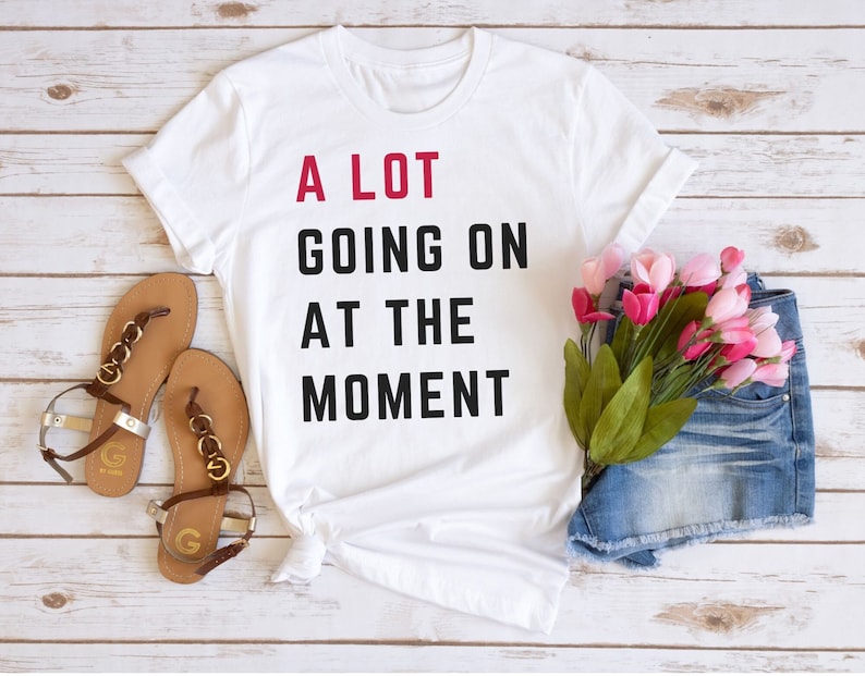 Pode incluir: Camiseta branca com o texto "A LOT GOING ON AT THE MOMENT" em preto e vermelho. A imagem inclui sand&aacute;lias marrons, shorts jeans e flores cor-de-rosa, em um fundo de madeira clara.