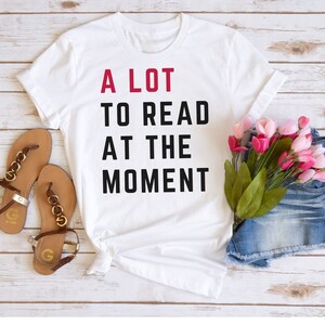 May include: White t-shirt with black text that reads "A LOT TO READ AT THE MOMENT" in a stacked format. The text "A LOT" is in red. The shirt is laid flat on a white wooden surface with a pair of brown sandals and a pair of denim shorts with pink flowers.