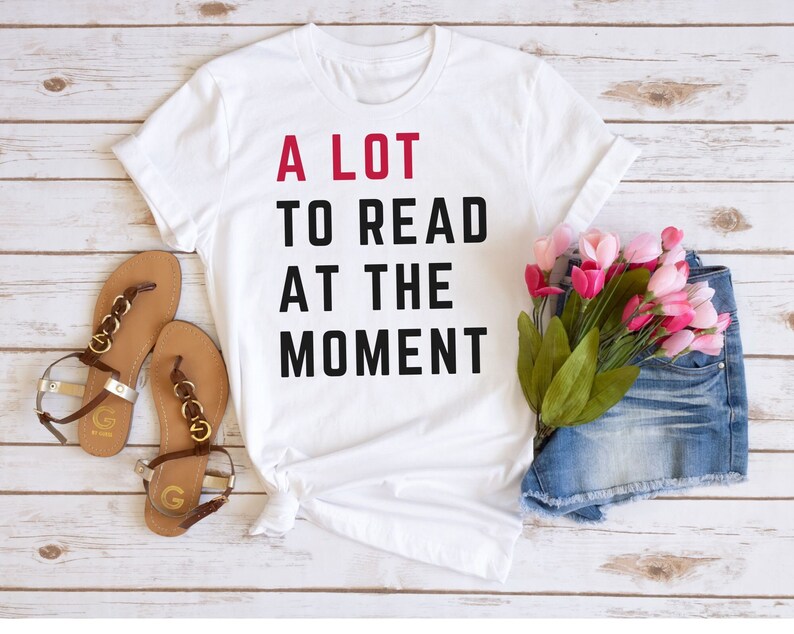 May include: White t-shirt with black text that reads "A LOT TO READ AT THE MOMENT" in a stacked format. The text "A LOT" is in red. The shirt is laid flat on a white wooden surface with a pair of brown sandals and a pair of denim shorts with pink flowers.