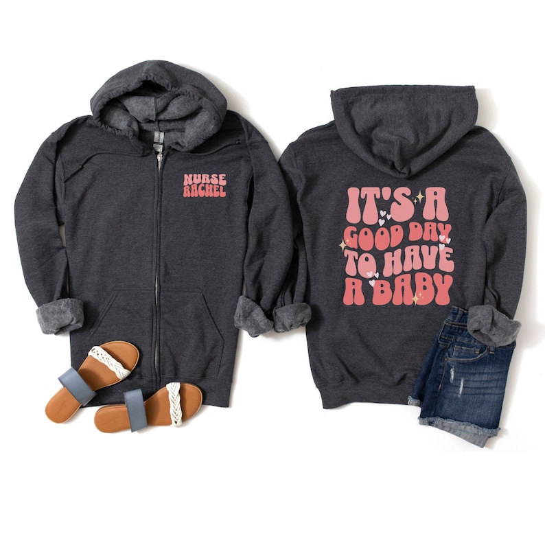 Puede incluir: Sudadera con capucha gris oscuro con cremallera con el texto "Nurse Rachel" en la parte delantera y "It's a good day to have a baby" en la parte trasera. La sudadera con capucha se muestra con sandalias azules y pantalones cortos de mezclilla.