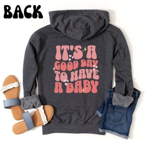 Puede incluir: Sudadera con capucha gris oscuro con el texto "IT'S A GOOD DAY TO HAVE A BABY" en letras rosas y rojas. La sudadera se combina con pantalones cortos de mezclilla azules y sandalias con correas trenzadas blancas.