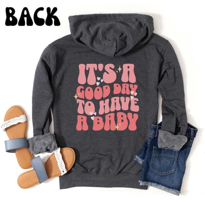 Puede incluir: Sudadera con capucha gris oscuro con el texto "IT'S A GOOD DAY TO HAVE A BABY" en letras rosas y rojas. La sudadera se combina con pantalones cortos de mezclilla azules y sandalias con correas trenzadas blancas.