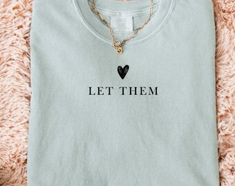 May include: A light green sweatshirt with the text "LET THEM" and a black heart graphic. A gold chain necklace with a small pendant is draped over the shirt. The sweatshirt is folded on a fluffy, peach-colored surface.
