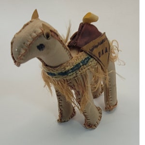 May include: A tan stuffed camel toy with a brown saddle and decorative fringe. The camel has stitched details, a blue and green woven band, and the letters "UAA" on the saddle. The camel is on a white background.