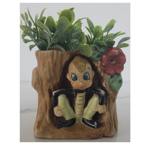 May include: A vintage ceramic planter shaped like a tree trunk, featuring a seated figure with a black jacket and light green pants. The planter is filled with green artificial foliage and a red flower.
