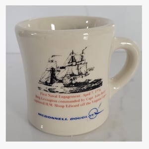 May include: Cream-colored ceramic mug featuring a black and white illustration of two sailing ships. The mug has a curved handle and text that reads "First Naval Engagement, April 7, 1776" and "McDonnell Douglas."
