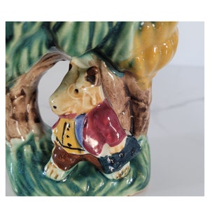 May include: A ceramic figurine depicting a whimsical animal character in a colorful outfit, set against a backdrop of stylized foliage. The figure wears a yellow vest, red coat, and carries a satchel. The base is a mix of green and brown.