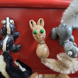 May include: Three whimsical animal figurines: a skunk, a rabbit, and a squirrel. The skunk is black and white, the rabbit is tan with green eyes, and the squirrel is gray. Each has a fluffy tail and is set against a red background.