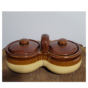 May include: A two-section ceramic condiment server with a handle. The server has two round compartments with matching lids. The top and lids are brown, while the bottom section is a light beige color. The server is on a wooden surface.