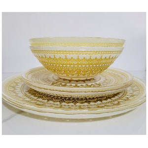 May include: A stack of vintage yellow and cream-colored dinnerware. The set includes two bowls and several plates. The bowls and plates have a decorative, embossed pattern. The dinnerware is arranged on a white surface.