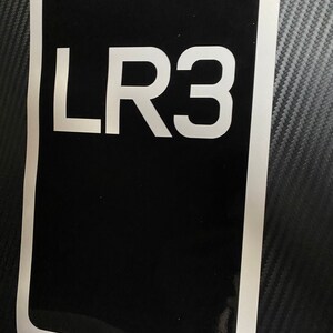 May include: A black rectangular sticker with the white text "LR3" in the centre. The sticker has a white border. The background is a dark grey, textured surface.