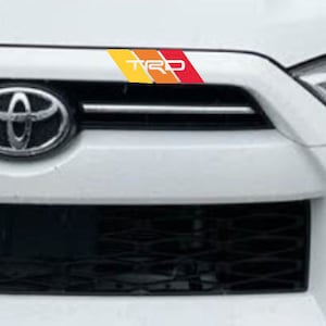 May include: A close-up of a white car's front grill with a colourful TRD sticker. The sticker has yellow, orange, and red stripes with the letters "TRD" in white. The Toyota logo is visible.