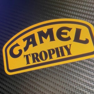 Puede incluir: Una pegatina ovalada amarilla y negra con el texto "CAMEL TROPHY" en una fuente estilizada.