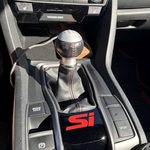 May include: A manual transmission gear shifter with a round silver and black knob featuring a six-speed shift pattern. The black leather boot has red stitching, and the center console displays a red Si logo, a parking brake button, and a sport mode button.