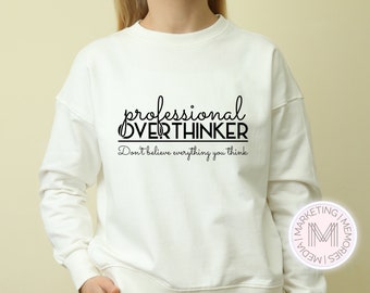 Op de afbeelding: Witte trui met zwarte tekst die "professional overthinker don't believe everything you think" luidt.