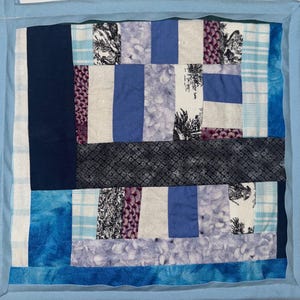 May include: A square, handmade quilt featuring a patchwork design in shades of blue, white, and purple. The quilt incorporates various fabric patterns, including floral prints, stripes, and solid colors. A light blue border frames the quilt.