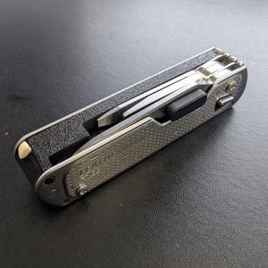 May include: A silver multi-tool with the brand name "LEATHERMAN" etched on the side. The tool is partially open, revealing a file and other implements. The handle has a textured, dotted pattern. The tool is resting on a black surface.