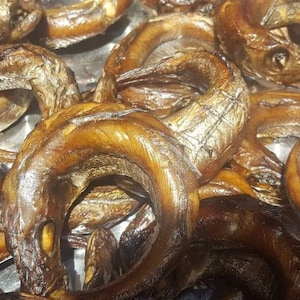 May include: A close-up of a pile of smoked fish. The fish are arranged in a circular pattern, with the tails pointing outwards. The fish are a dark brown color and have a shiny, oily surface.