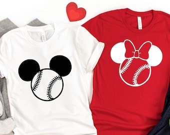 May include: Two white t-shirts with a baseball and Mickey Mouse ears design. The shirt on the left has black Mickey Mouse ears and a white baseball. The shirt on the right has white Mickey Mouse ears and a red baseball. The shirts are laid out on a white surface with a red heart and a pair of blue jeans.