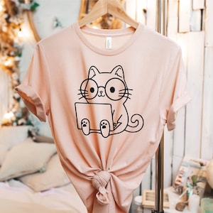 May include: A light pink t-shirt with a black and white illustration of a cat wearing glasses and using a laptop.