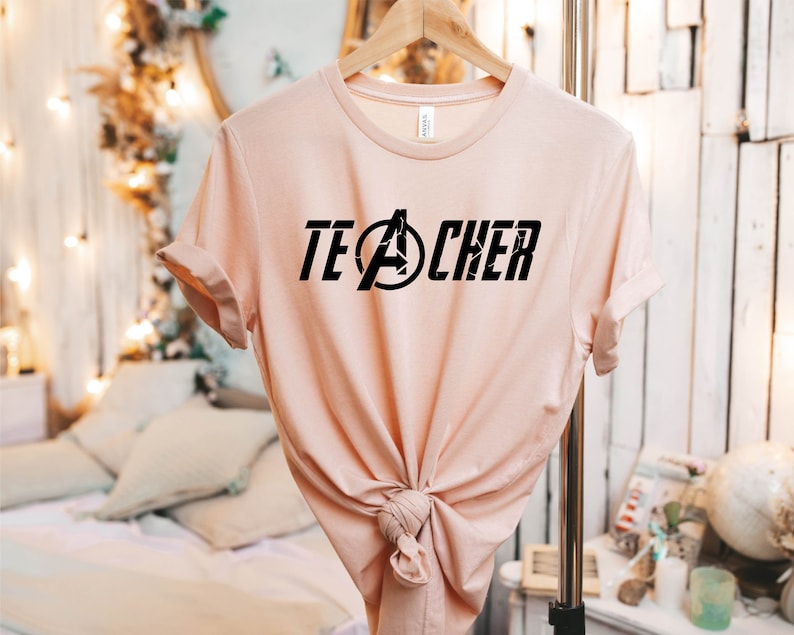 Op de afbeelding: Een lichtroze T-shirt met een zwarte afbeelding waarop "TEACHER" staat, waarbij de "A" is vervangen door het Avengers-logo.