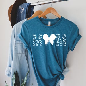 Puede incluir: Una camiseta azul turquesa con un gr&aacute;fico blanco de las letras "MOM" con una silueta de cabeza de Mickey Mouse en el centro. Las letras est&aacute;n rellenas con un patr&oacute;n de estampado de leopardo blanco.