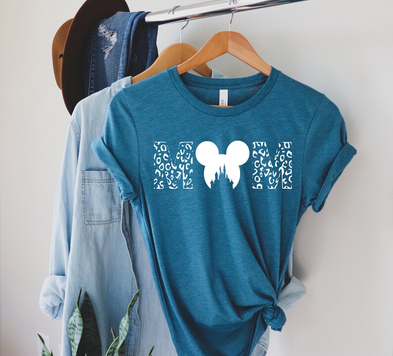 Puede incluir: Una camiseta azul turquesa con un gr&aacute;fico blanco de las letras "MOM" con una silueta de cabeza de Mickey Mouse en el centro. Las letras est&aacute;n rellenas con un patr&oacute;n de estampado de leopardo blanco.