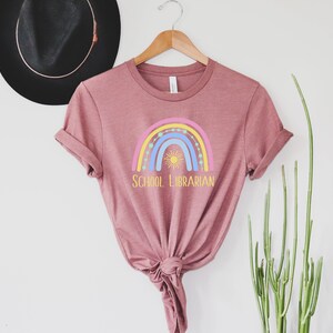 May include: A mauve t-shirt with a rainbow and sun graphic and the text "School Librarian".